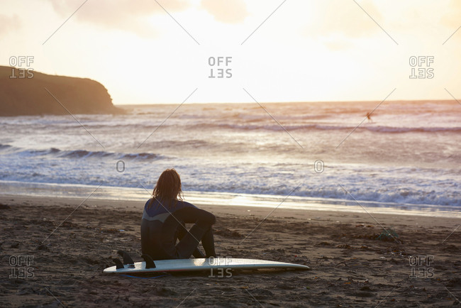 Young male surfer sitting looking out from beach, Devon, England, UK