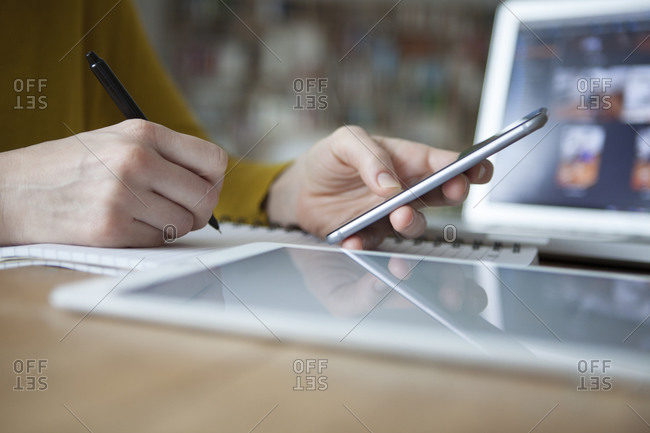 Woman at table using smartphone and writing in notebook