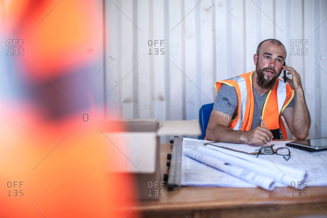 Construction worker sitting at desk talking on smartphone in portable cabin