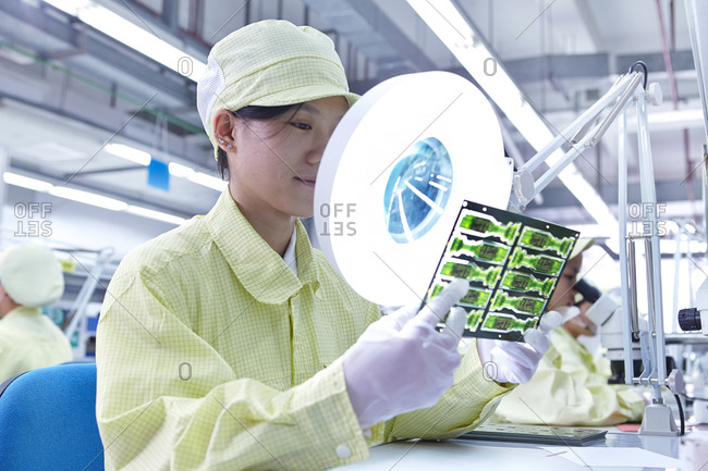 Female worker using magnifying glass at quality check station for a factory producing flexible electronic circuit boards