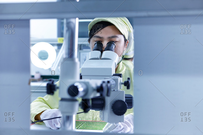 Young woman at microscope at quality check station at factory producing flexible electronic circuit boards