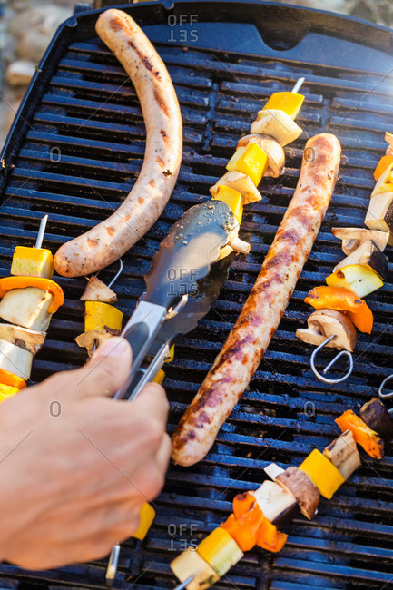 Person using tongs to turn sausages and kebabs on barbecue griddle