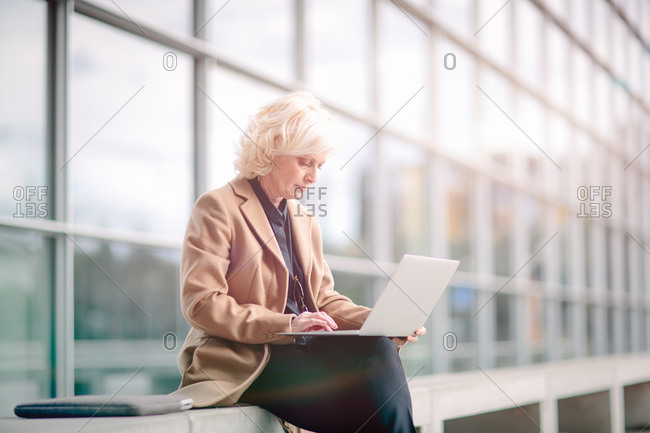 Mature businesswoman, sitting on bench, outside building, using laptop