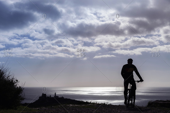 Silhouetted rear view of male mountain biker on coastal path at dusk, Cagliari, Sardinia, Italy