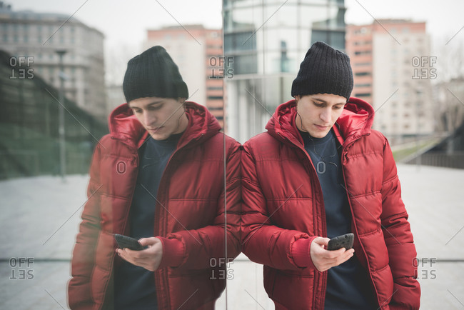 Mirror image of young man leaning against office building reading smartphone texts
