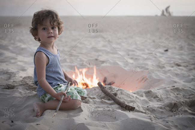 Boy sitting next to camp fire on beach and family in the background, Lit-et-Mixe, Aquitaine, France