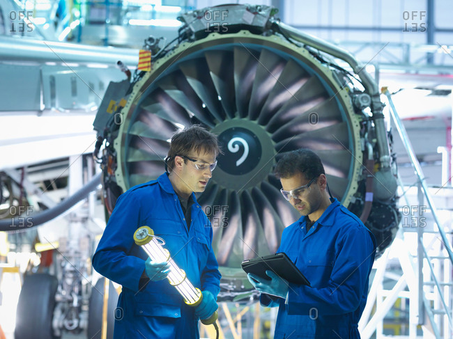 Aircraft engineers discussing maintenance details on digital tablet in front of jet engine in aircraft maintenance factory