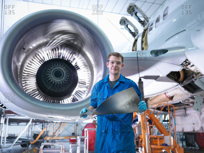 Portrait of a young engineer holding jet engine turbine blade in aircraft maintenance factory