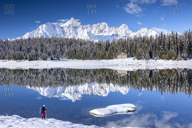 Hiker admires the snowy peaks and woods reflected in Lake Pali Malenco Valley Valtellina Lombardy Italy Europe