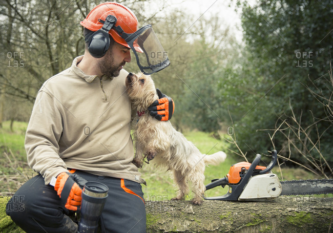 A forester sitting on a log in woodland wearing a helmet and ear defenders, hugging a small dog