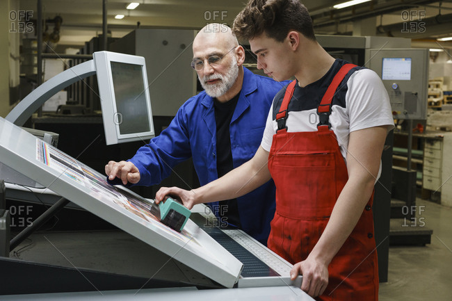 Mature man and coworker doing quality check of printout with machinery at printing press