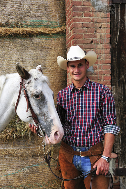 Portrait of young man in cowboy gear with horse outside stable