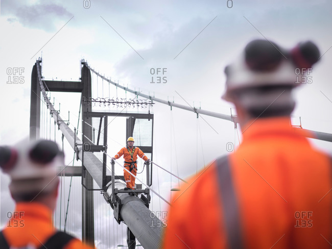 Bridge workers watching coworker on cable of suspension bridge