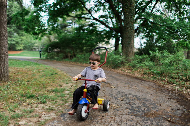 Boy wearing sunglasses riding a tricycle down a tree lined path