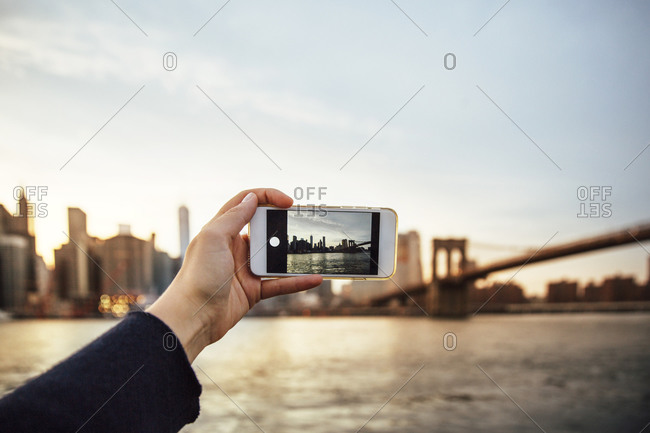 Cropped hand of woman photographing skyline and Brooklyn Bridge at dusk