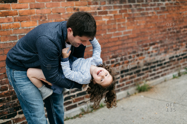 Father holds young daughter upside down outside brick building