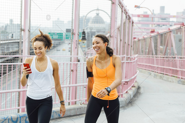 Two woman in workout apparel laughing and interacting as they walk along a bridge