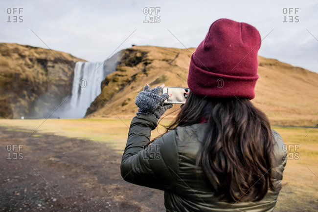 Female tourist taking smartphone photographs of waterfall  at Skogafoss, Iceland