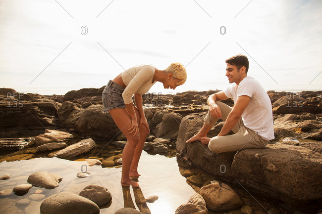 Young woman standing in rock pool, man sitting on rocks observing
