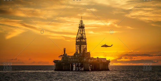 Helicopter flying over oil rig in sea against sky during sunset