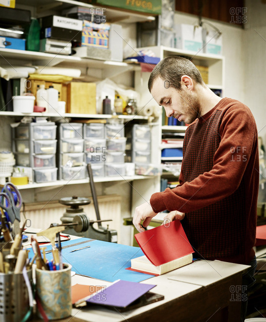 A man working in a book binding workshop, creating a red cover for freshly stitched pages