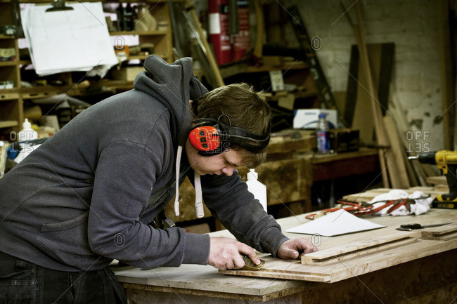 A man working in a furniture maker\'s workshop wearing ear defenders and using a sharp chisel on wood