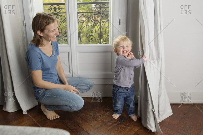 Smiling toddler playing peek-a-boo behind the curtain while his mother watching him