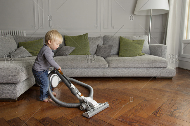 Toddler vacuuming a wooden floor by a gray couch