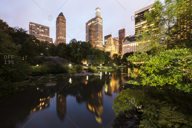 Lake amidst trees at Central Park by illuminated buildings at dusk