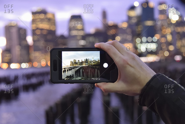 Person holding a smart phone, taking a photograph of a city skyline in the evening