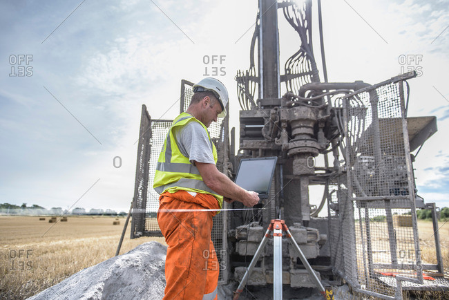 Worker using laptop to survey drilled hole made by drilling rig in field