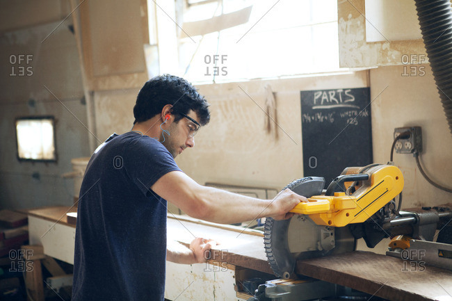 Side view of male carpenter cutting wooden plank with electric saw in workshop