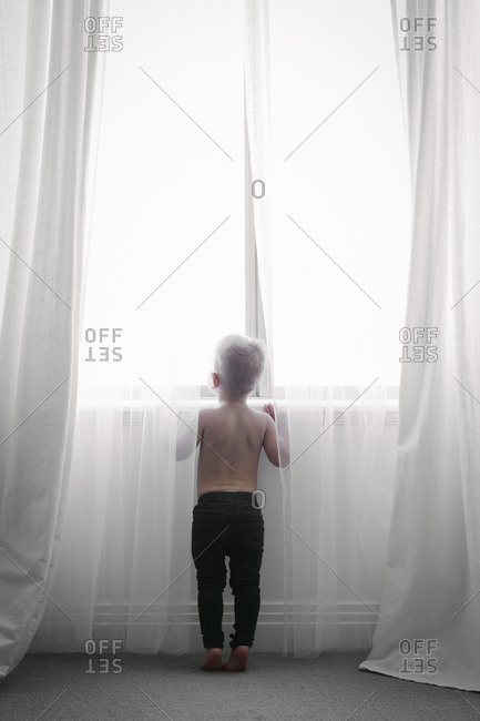 Back view of a child standing at a window looking out through the net curtains
