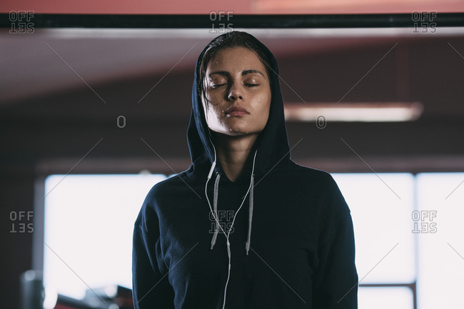 Exhausted woman wearing hooded shirt standing with eyes closed at gym
