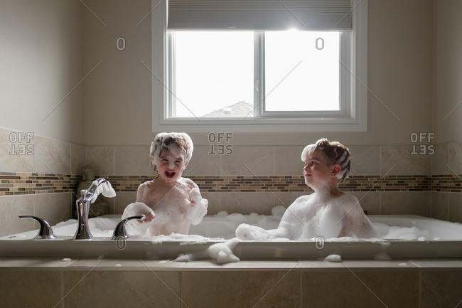 Two young children covered in soap bubbles play in bathtub