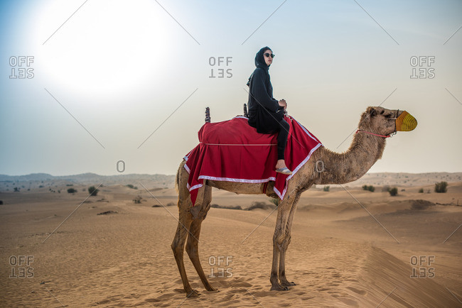 Young woman wearing traditional middle eastern clothes riding camel in desert