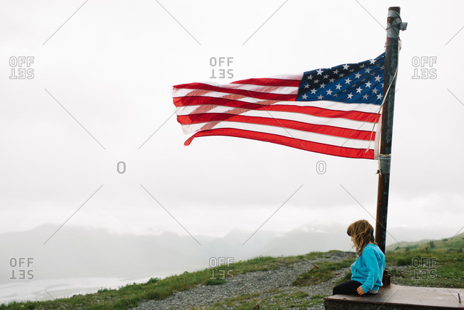 Girl sitting below an American flag on a mountain summit