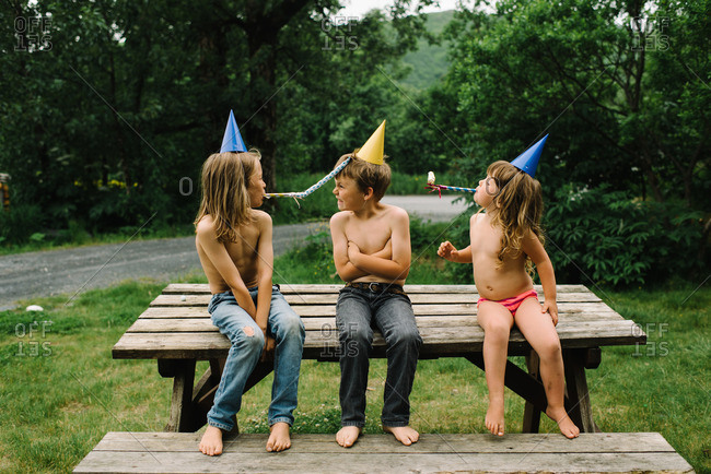 Boy sitting on a picnic table while brother and sister blow party noisemakers