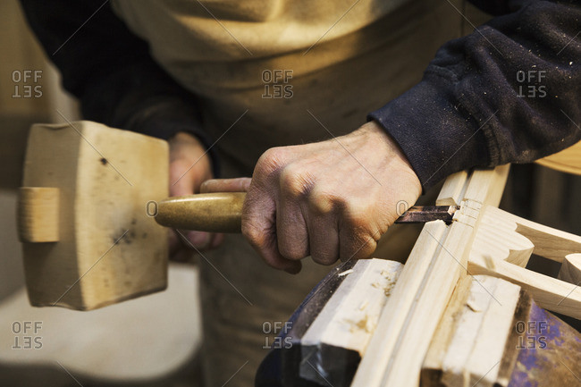 Close up of a man standing at a work bench in a carpentry workshop, working on a wooden chair with a wooden mallet and chisel