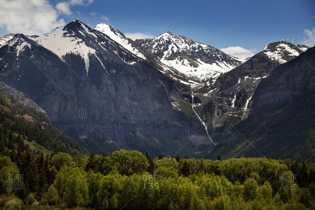 Snowcapped Telluride, Colorado mountains with a waterfall and stream tumbling down a steep mountainside and sunlit trees in the foreground