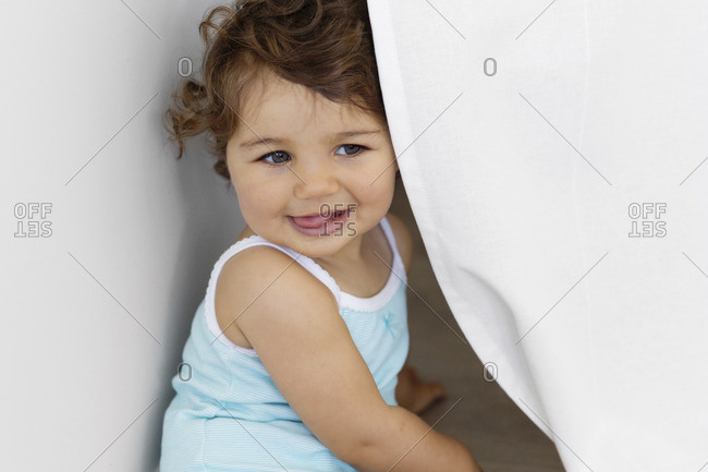 Portrait of baby girl playing hide and seek behind a white curtain with her tongue sticking out