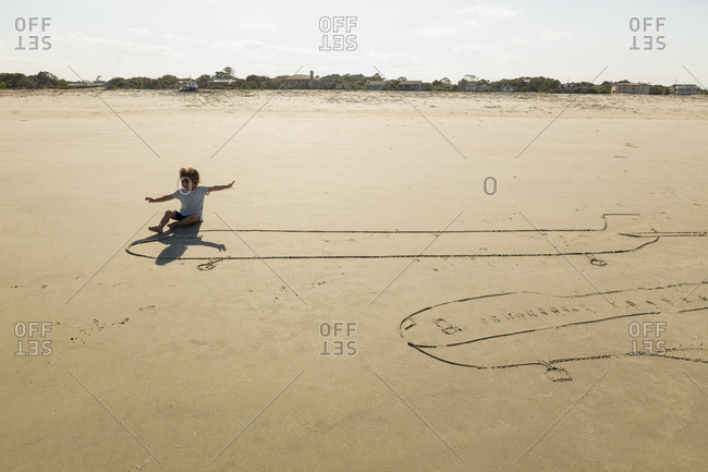 Boy sitting on plane drawing in the sand with his arms spread