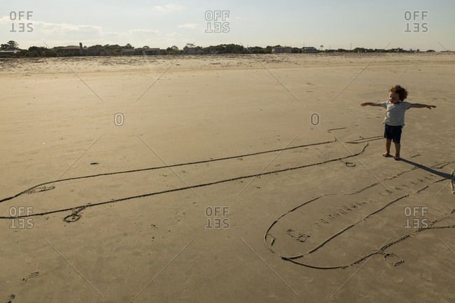 Boy standing by plane drawings in the sand with his arms spread