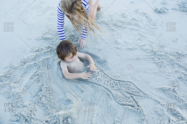 Girl burying little boy in sand with mermaid fin drawing