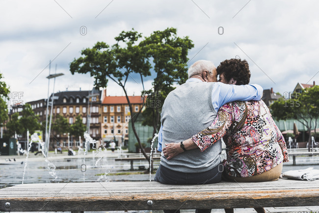 Germany- Mannheim- back view of kissing senior couple sitting on a bench hugging each other
