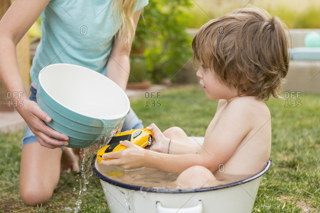 Toddler boy cooling off in a water bucket while his older sister pours water from a bowl