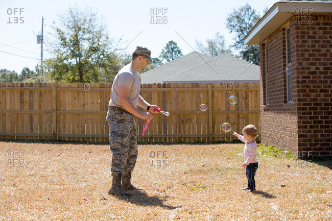 Young male soldier blowing bubbles with toddler daughter in garden at air force military base