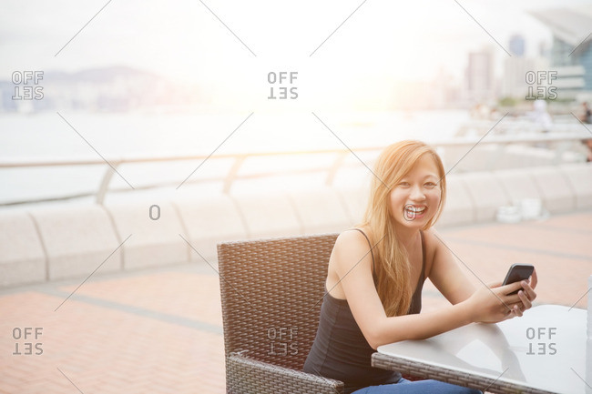 Young woman sitting beside harbor, using smartphone, Hong Kong, China