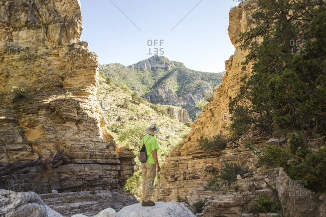 A man in hiking clothes stands on a rock outcrop looking through a V-shaped slot in a desert canyon