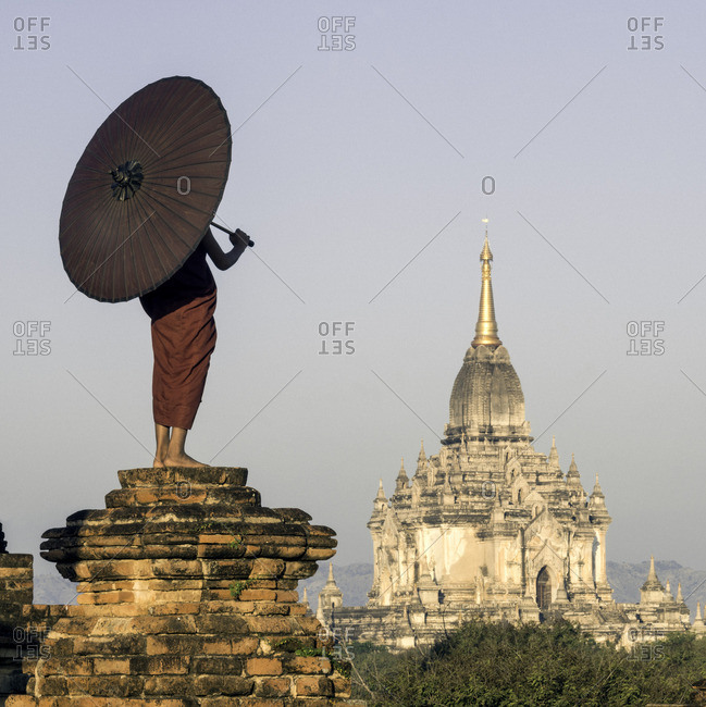 Young Buddhist monk standing on temple top holding a parasol in front of an ancient temple